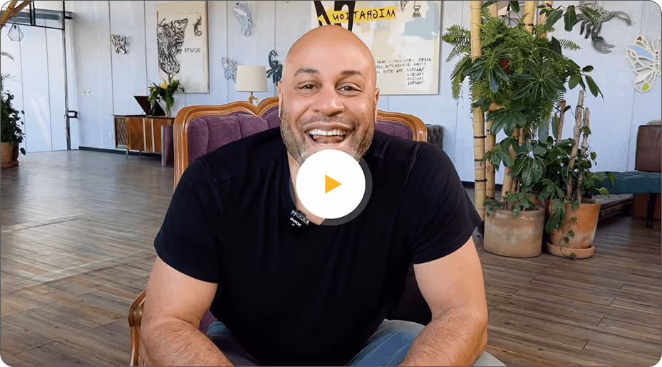 Smiling bald man wearing a black t-shirt sitting in a purple chair in a room with plants and wall decorations.