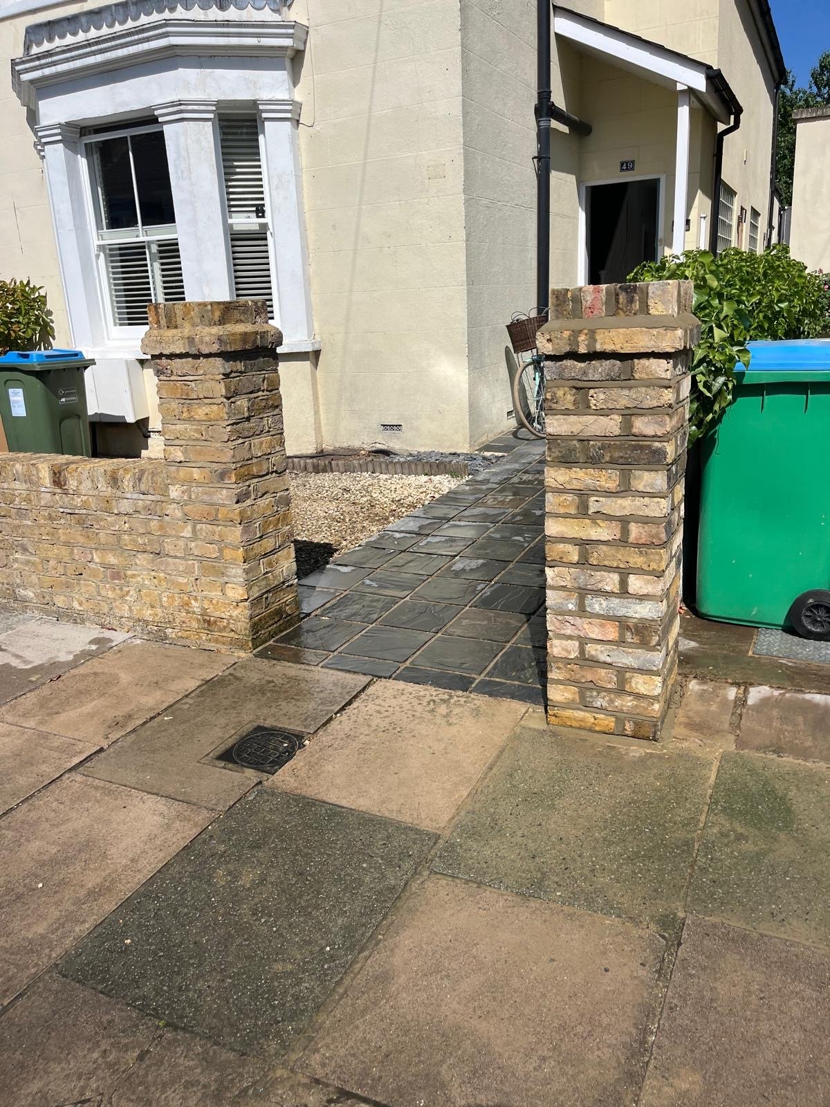 Entrance to a beige house with a tiled pathway between two brick pillars and green and blue trash bins nearby.