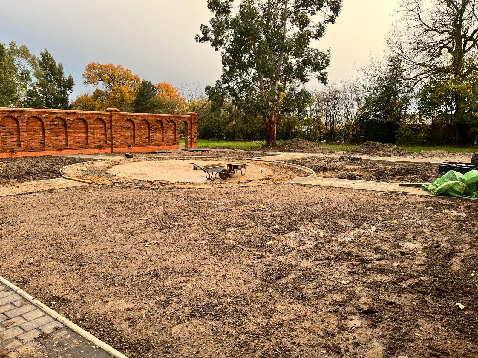 Construction site for a garden or outdoor area with bare soil, two wheelbarrows on a circular paved section, a red brick wall with arched openings, and trees in the background.