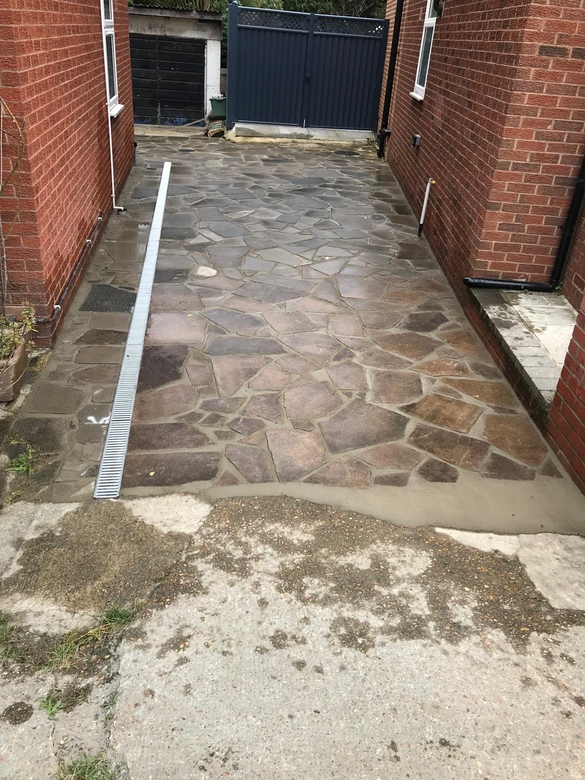 Newly paved stone driveway between two brick houses with a metal drainage grate on the left side and a closed dark blue gate at the end.