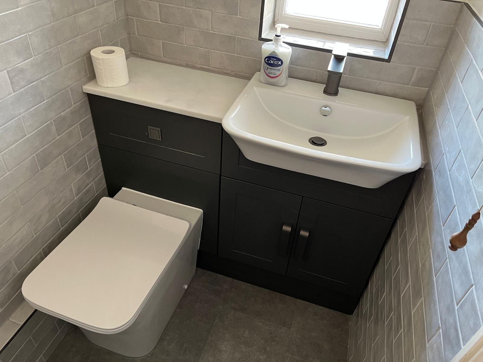 Small modern bathroom with white toilet, black vanity with white sink, gray tiled walls, and a roll of toilet paper on the counter.