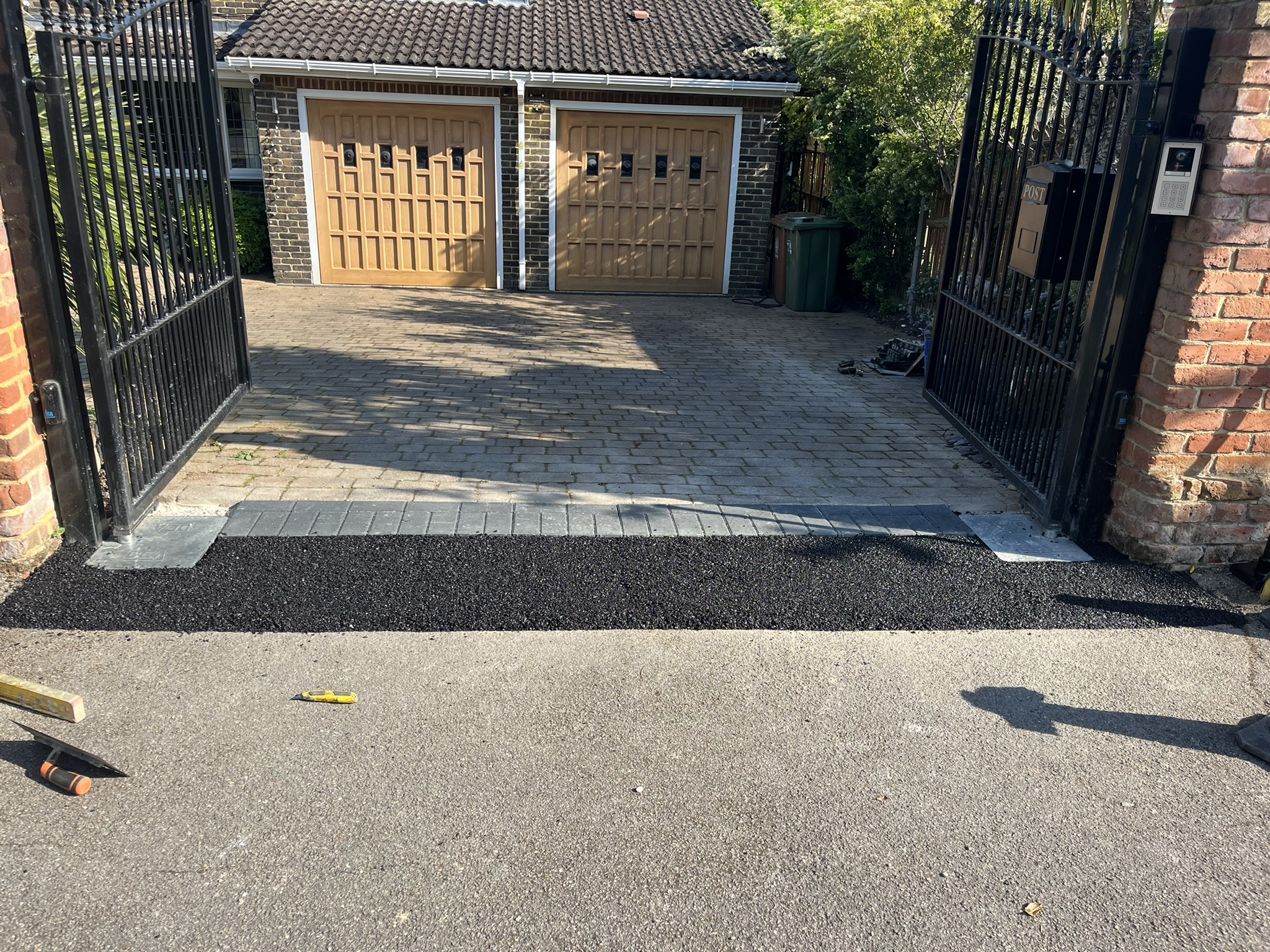 Open black metal gates leading to a brick driveway with double wooden garage doors and fresh black asphalt patch at the entrance.