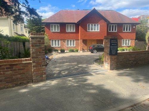 Two-story brick and red-shingle house with a paved driveway, a black car parked outside, and brick pillars at the entrance.