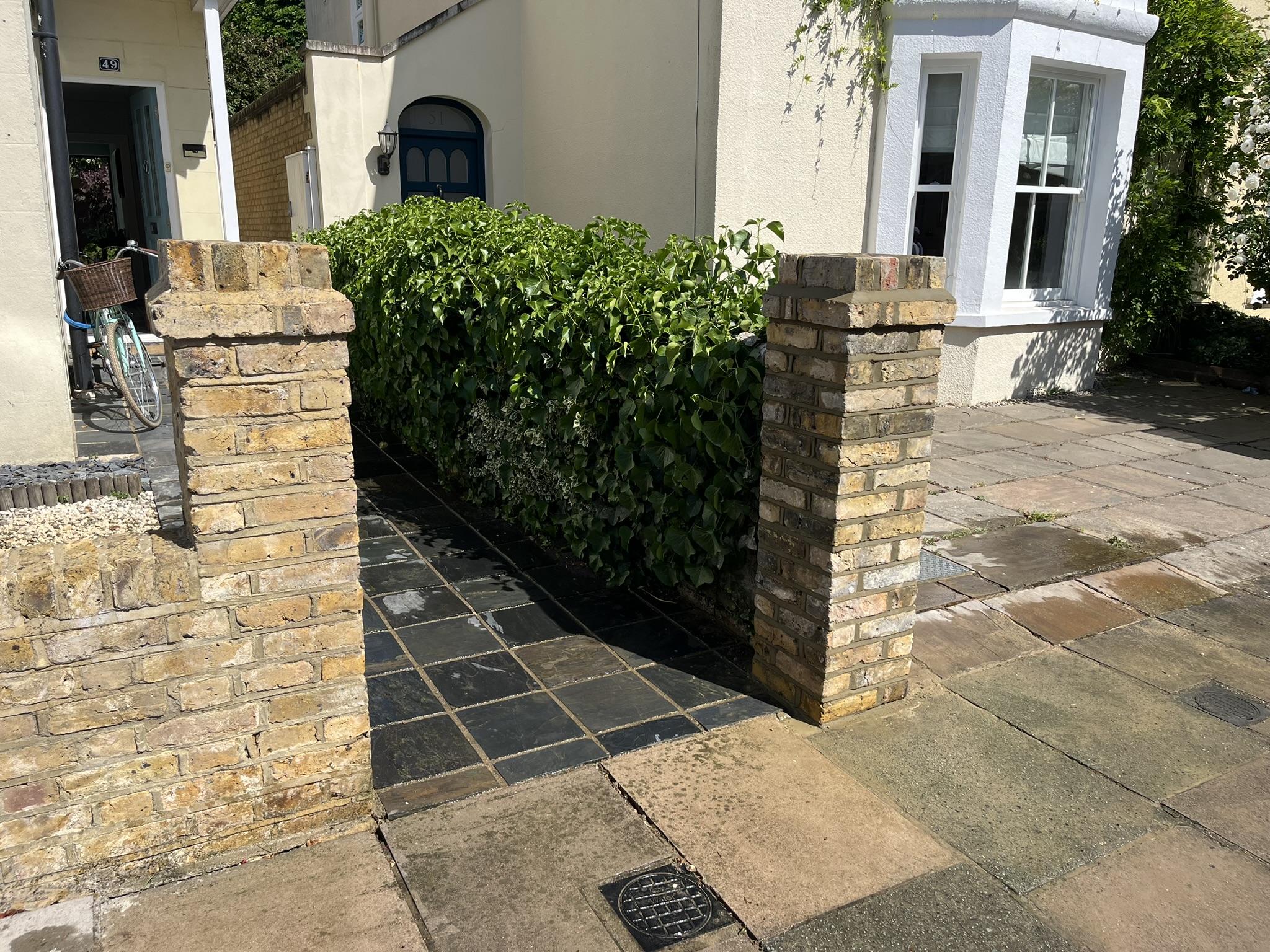 Brick pillars framing a tiled pathway leading to a house entrance with green bushes on the right side.