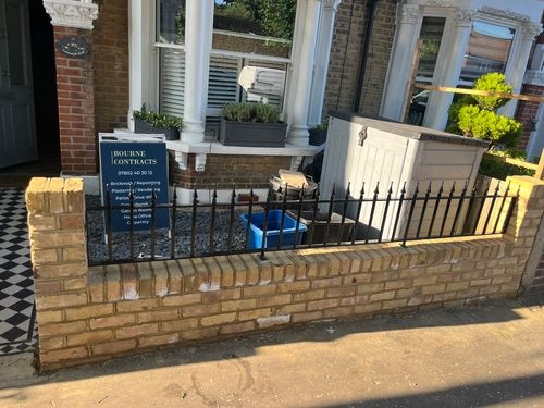 Front yard with a low brick wall and black metal fence, a blue sign for Bourne Contracts offering brickwork and carpentry, a gray storage box, and potted plants under a bay window.
