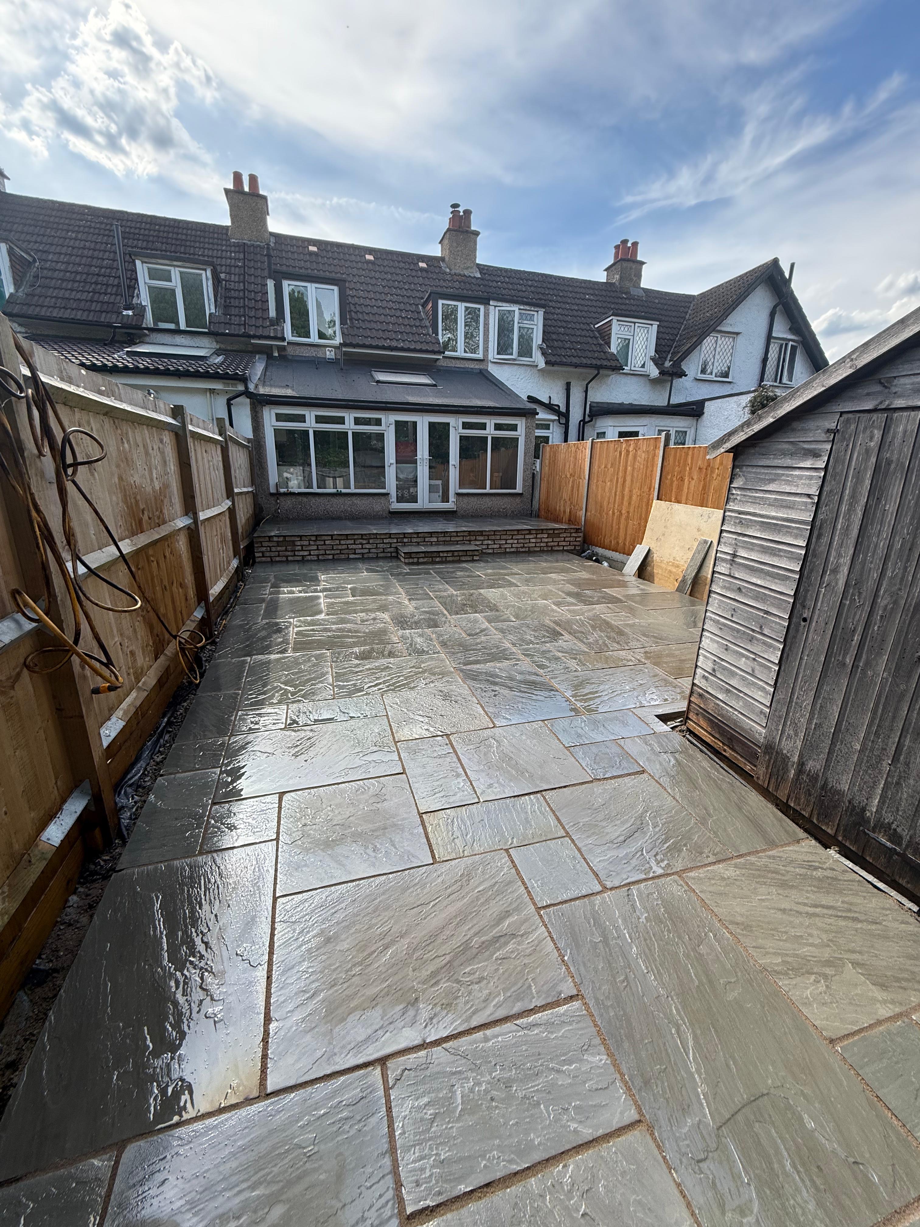 Backyard with newly installed wet stone tile patio, wooden shed on the right, and a two-story house with fenced sides under a partly cloudy sky.