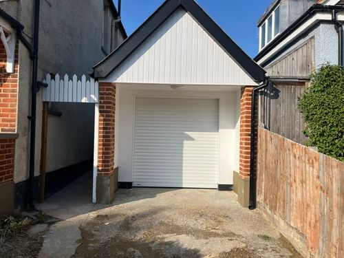 Small white garage with closed roll-up door, brick pillars, and a wooden fence on the right side.