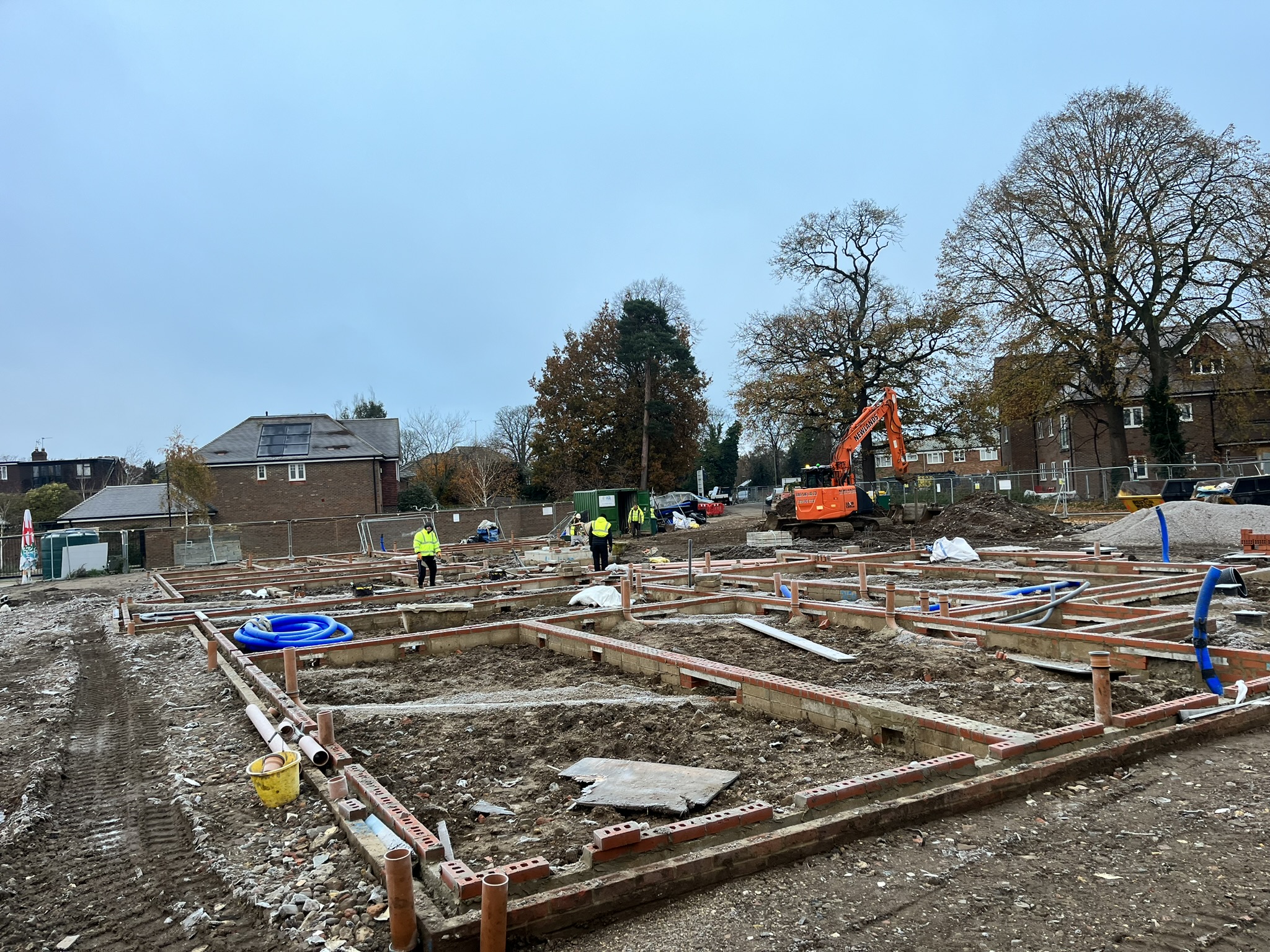 Construction site with foundations laid out, workers in high-visibility jackets, and an orange excavator amid trees and residential buildings.
