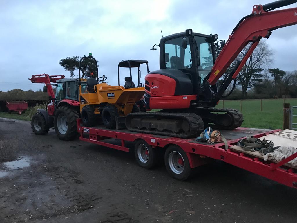 Red tractor towing a flatbed trailer carrying an orange mini dumper and a red mini excavator on a dirt road near green fields.