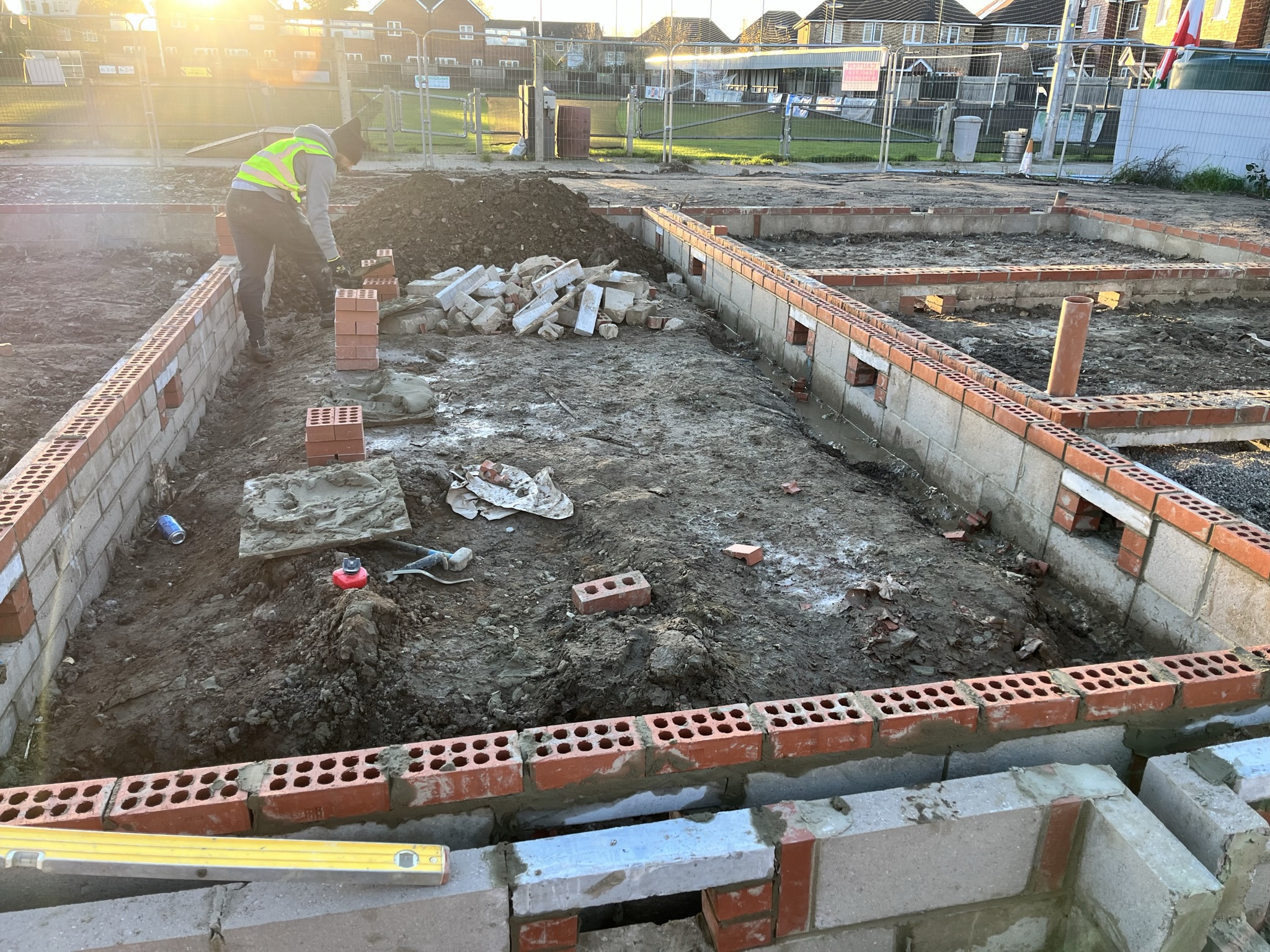 Construction site with a worker in a neon vest laying bricks on a foundation during sunset.