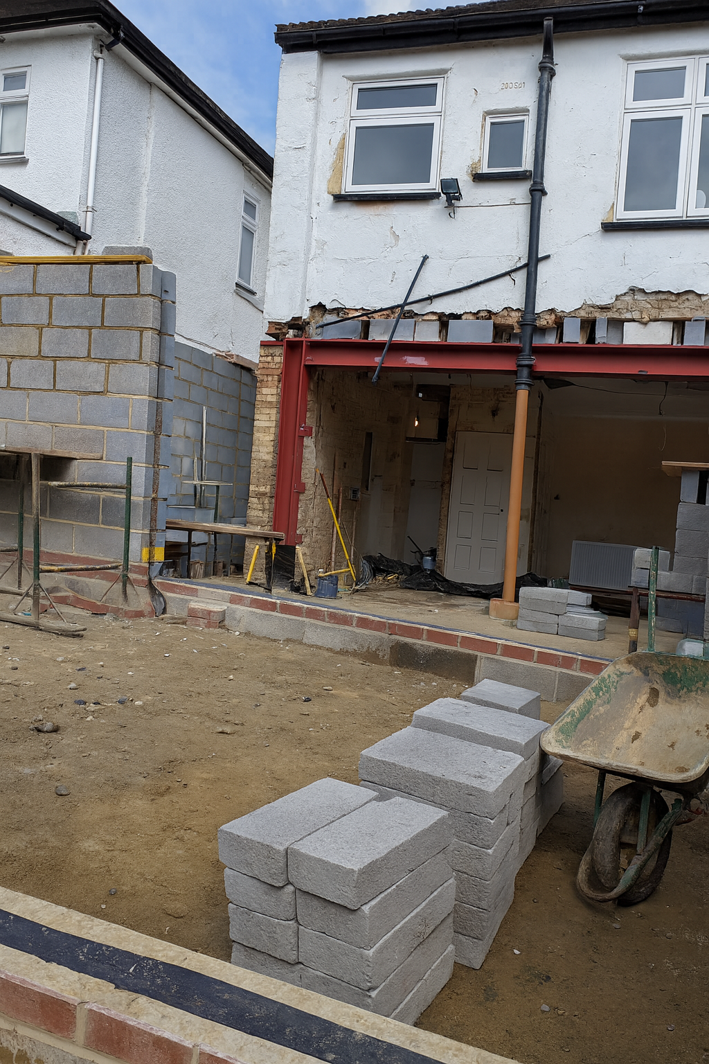 Construction site showing renovation of a two-story white house with bricks, a wheelbarrow, and scaffolding.