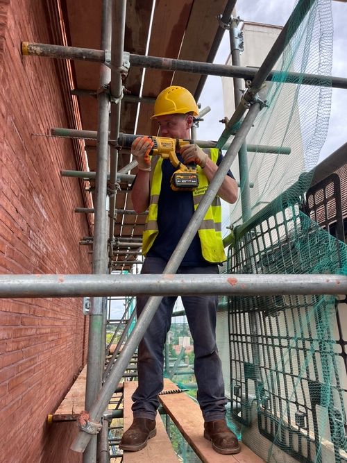 Construction worker wearing a yellow hard hat and reflective vest using a drill while standing on scaffolding next to a brick wall.