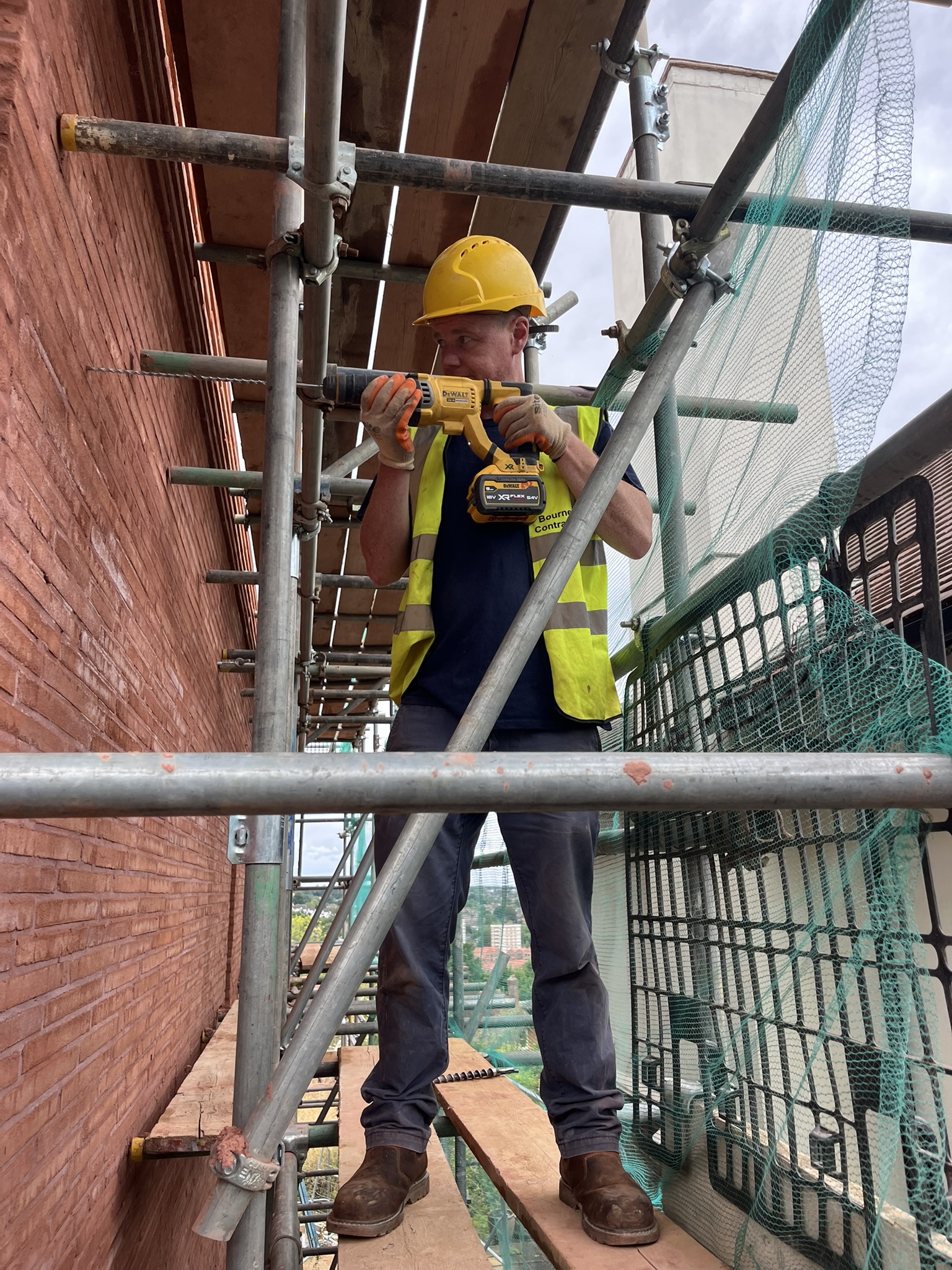 Construction worker wearing a yellow hard hat and reflective vest using a drill while standing on scaffolding next to a brick wall.