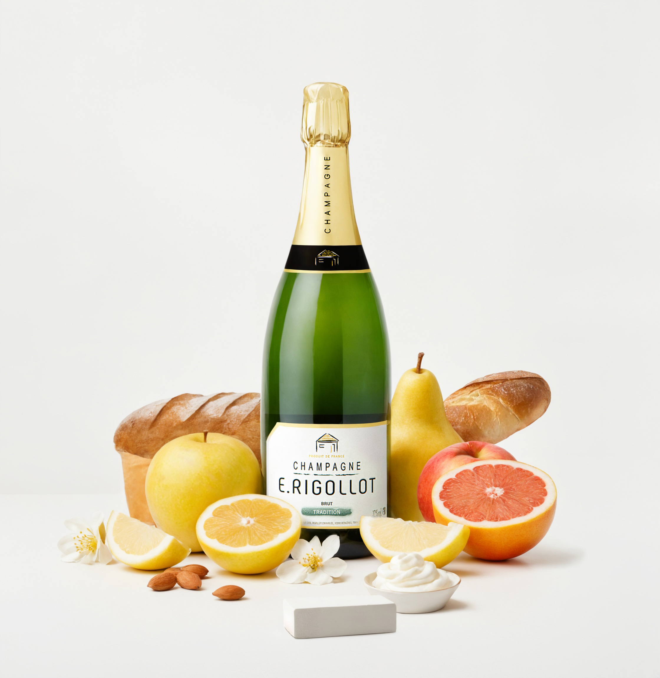 Bottle of E. Rigollot Champagne surrounded by lemon halves, whole lemon, apple, pear, grapefruit half, bread loaves, almonds, white flowers, small bowl of cream, and a white rectangular block on a white surface.