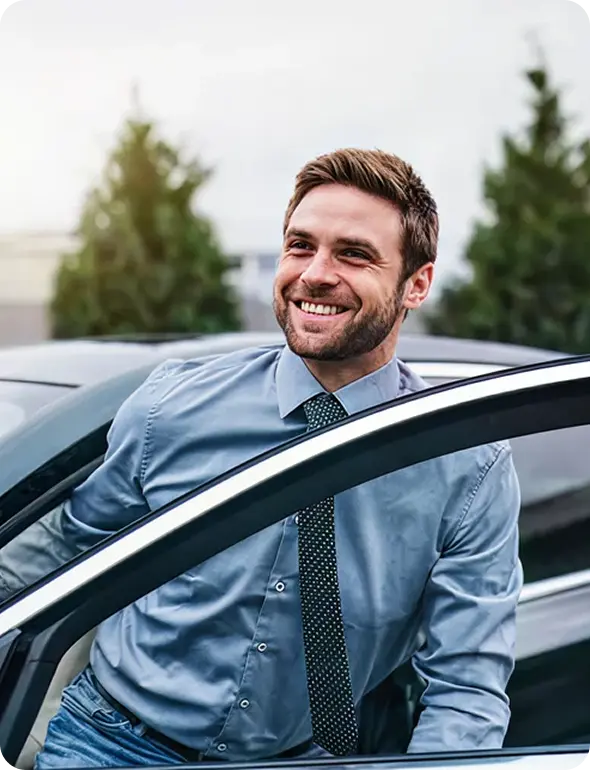 Smiling young man in a blue shirt and tie stepping out of a car with trees in the background.