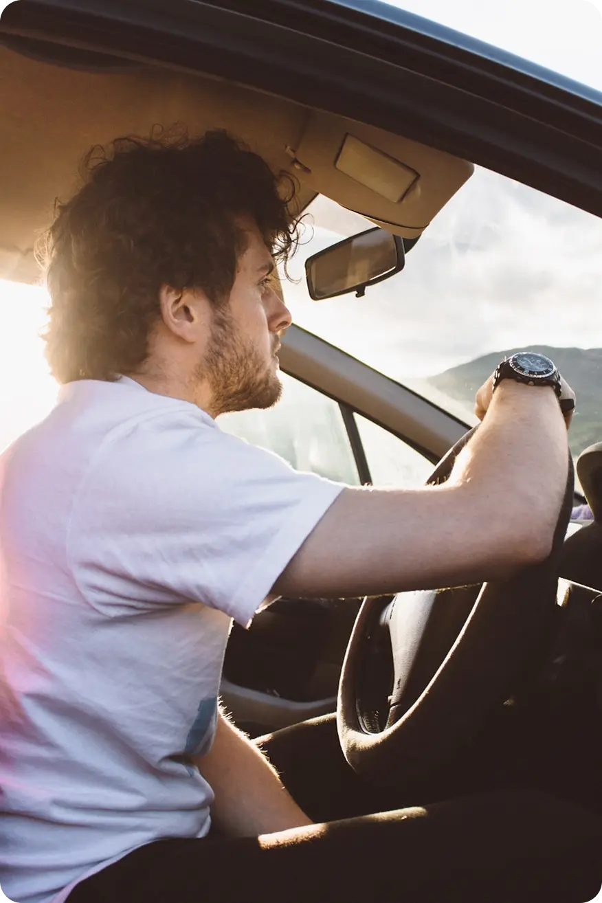 Man with curly hair and a beard driving a car, shown from the side in sunlight.