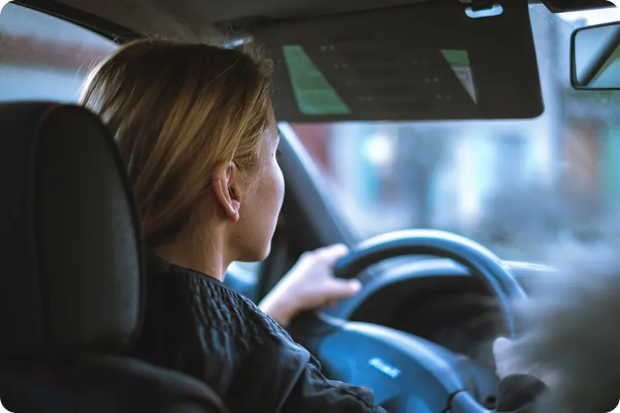 Woman with blonde hair driving a car, seen from behind the front passenger seat.