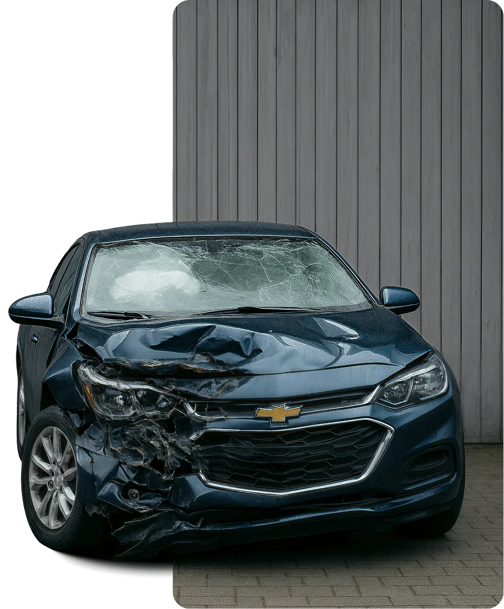 Dark blue Chevrolet car with heavy front-end damage and a shattered windshield, parked in front of a wooden fence.