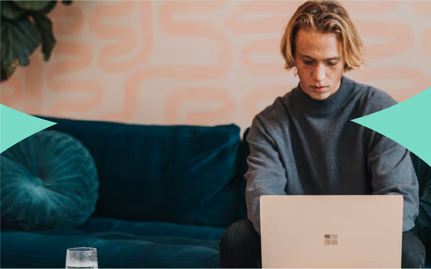 Young man with blonde hair sitting on a dark green couch, focused on a silver laptop with a blurred patterned wall background.