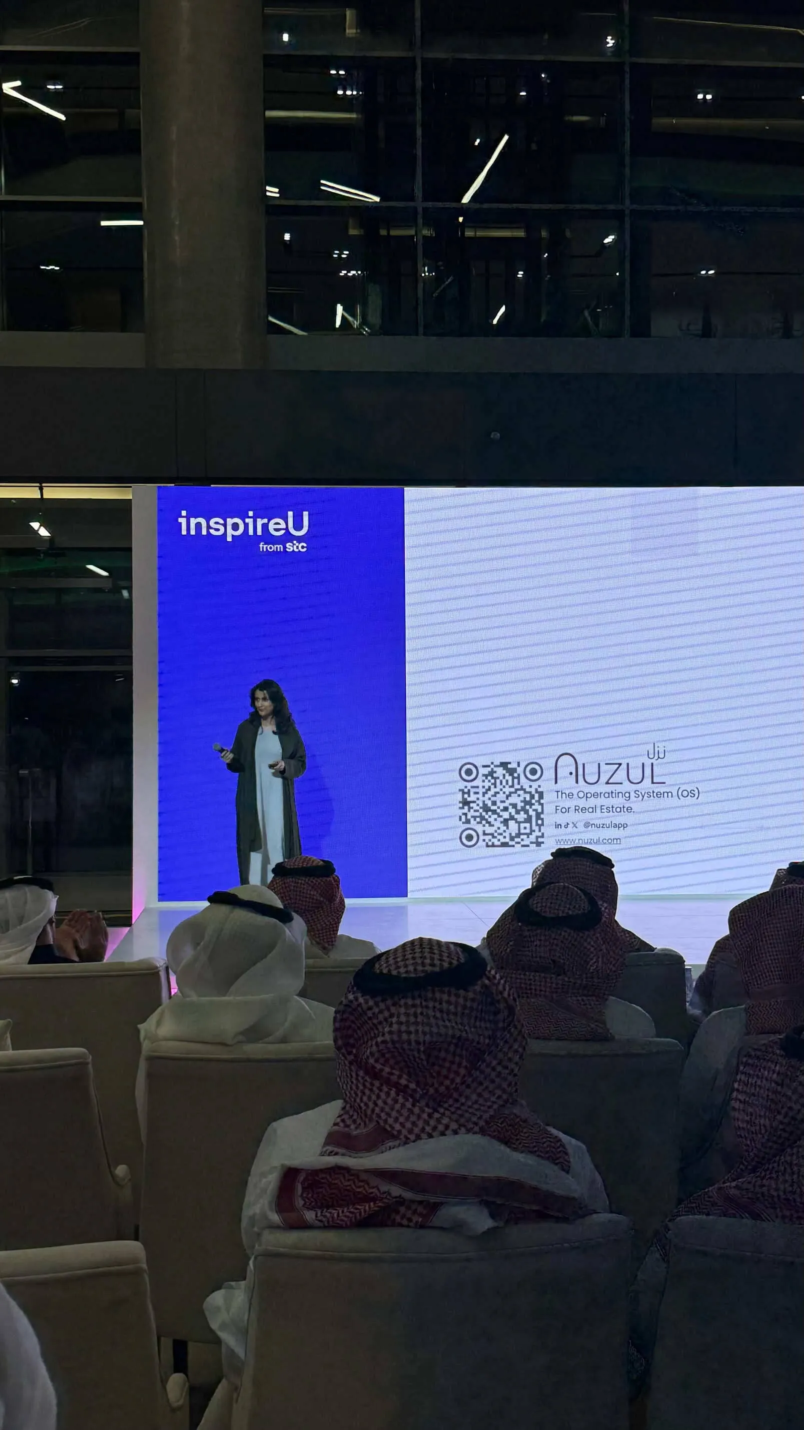 A woman giving a presentation on stage with a large screen behind her displaying 'inspireU from stc' and 'Nuzul The Operating System (OS) for Real Estate' along with a QR code, in front of an audience mostly wearing traditional Middle Eastern attire.