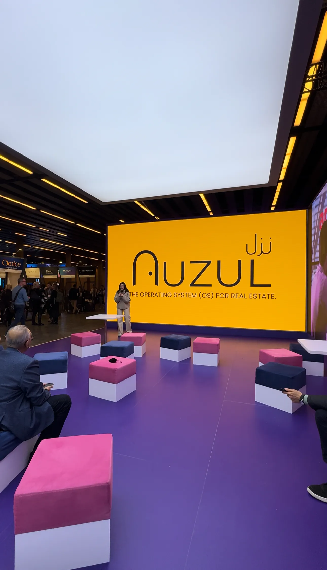Exhibition booth with purple floor, multicolored cushioned seats, and a large yellow display reading 'Auzul The Operating System (OS) for Real Estate,' with a woman standing nearby and attendees around.