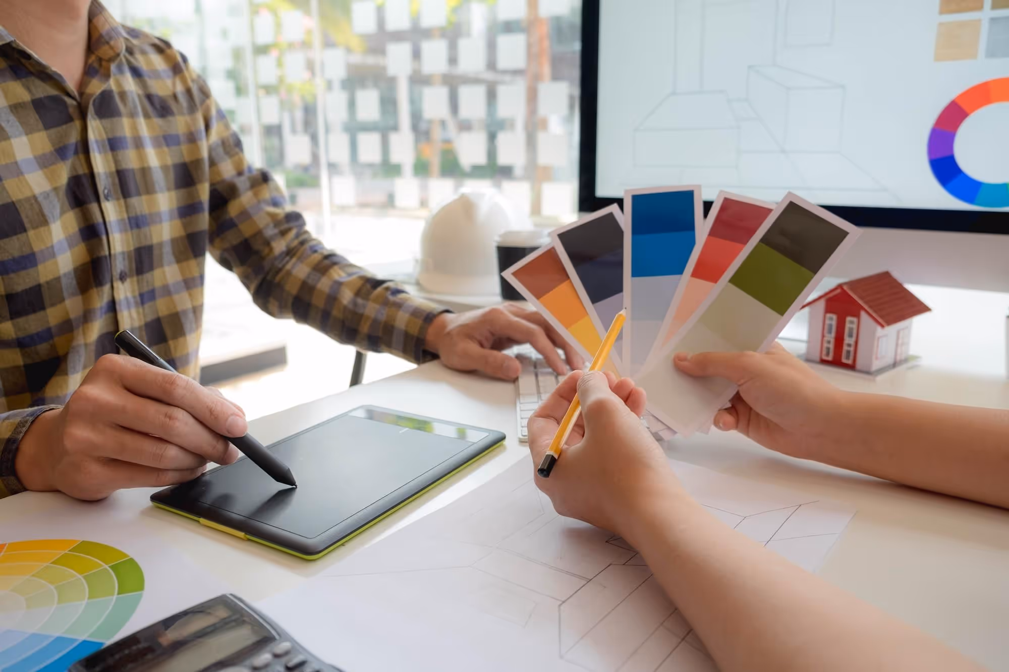 Two people discussing color swatches and architectural sketches with a digital drawing tablet, keyboard, and miniature house model on the table.