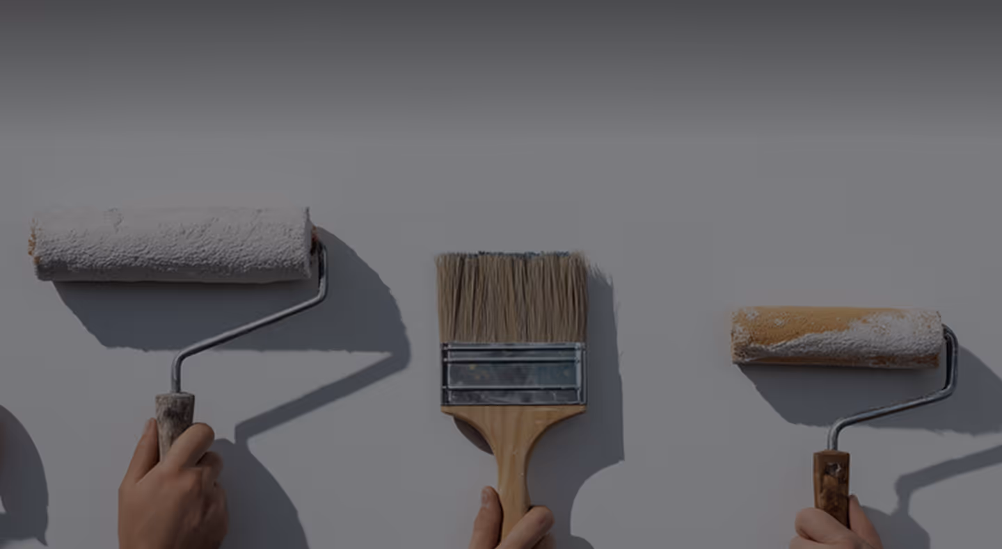 Three hands holding paintbrushes and rollers with white paint against a light gray wall.