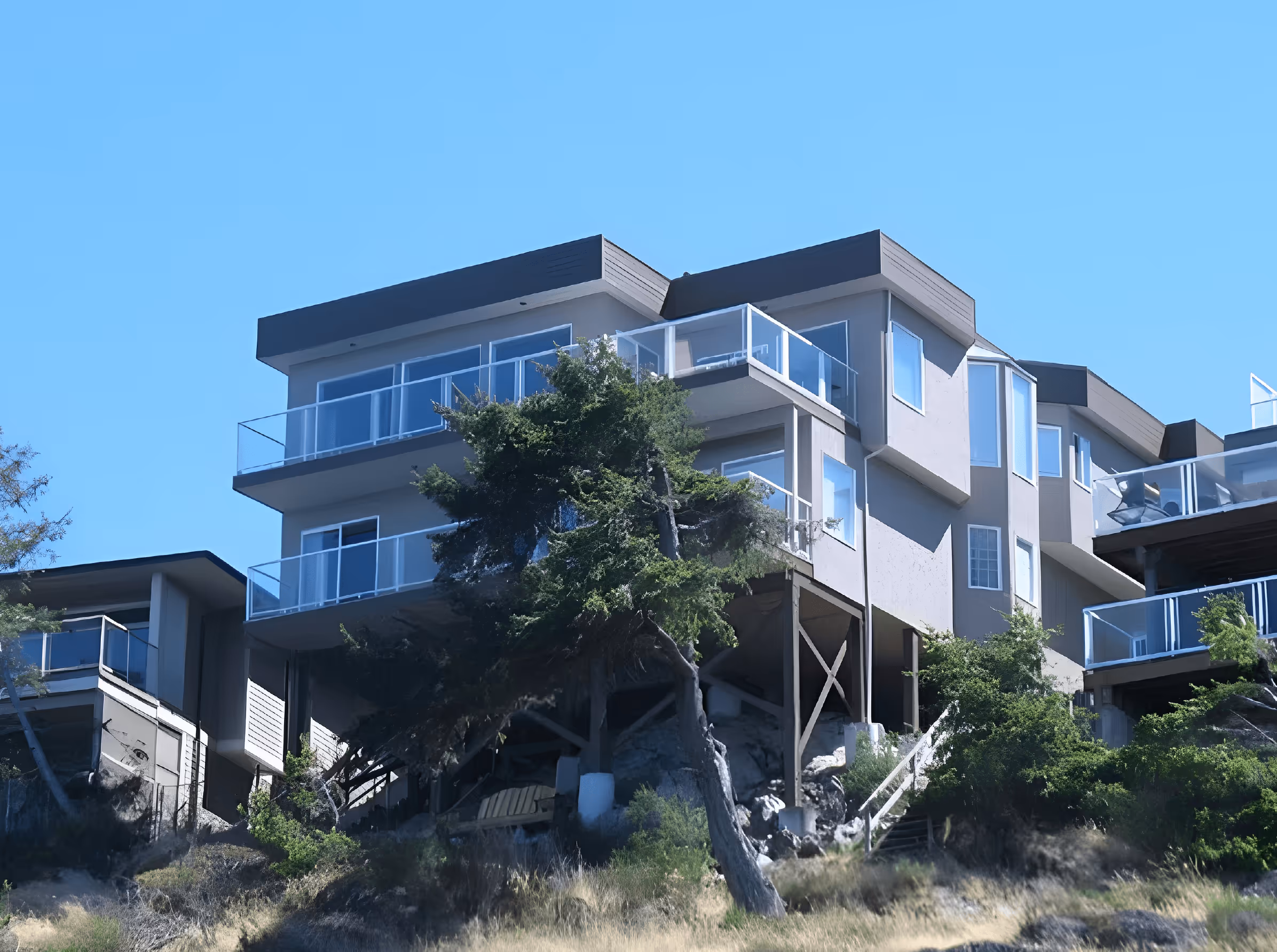 Modern multi-story house on stilts with glass railings perched on a hillside with trees and shrubs.