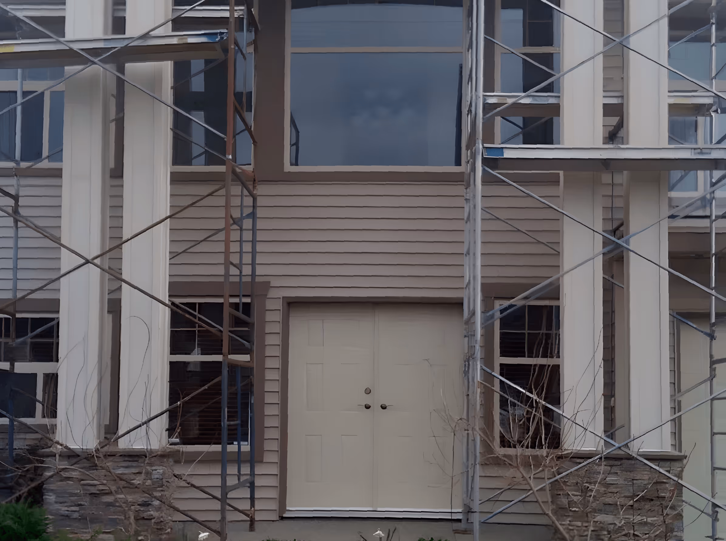 Front entrance of a beige house under construction with double doors, stone foundation, white columns, and scaffolding.