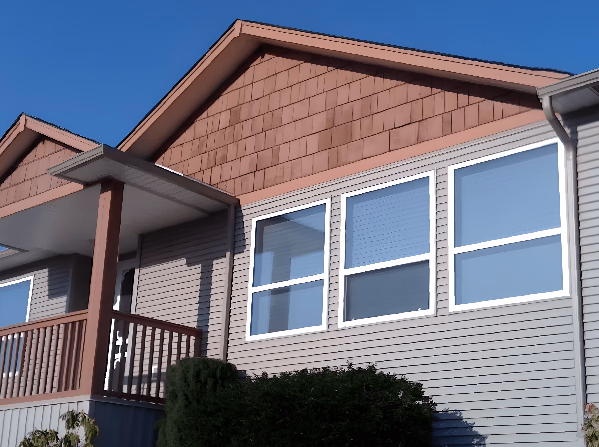 Close-up of a house exterior featuring beige siding, three large white-framed windows, a small porch with wooden railing, and a shrub in front under a clear blue sky.