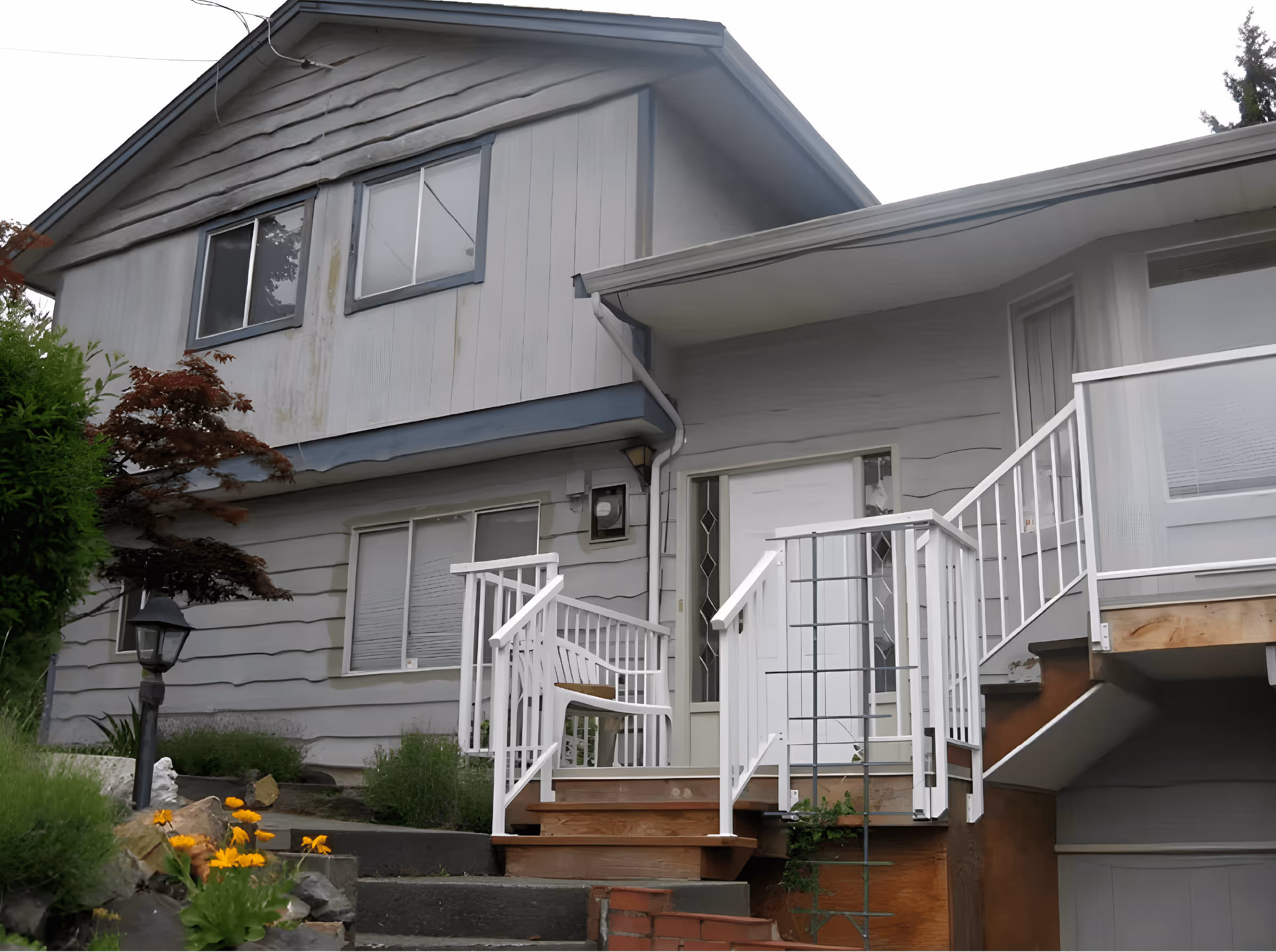 Gray two-story house with white railings and stairs leading to a front door, surrounded by green bushes and yellow flowers.
