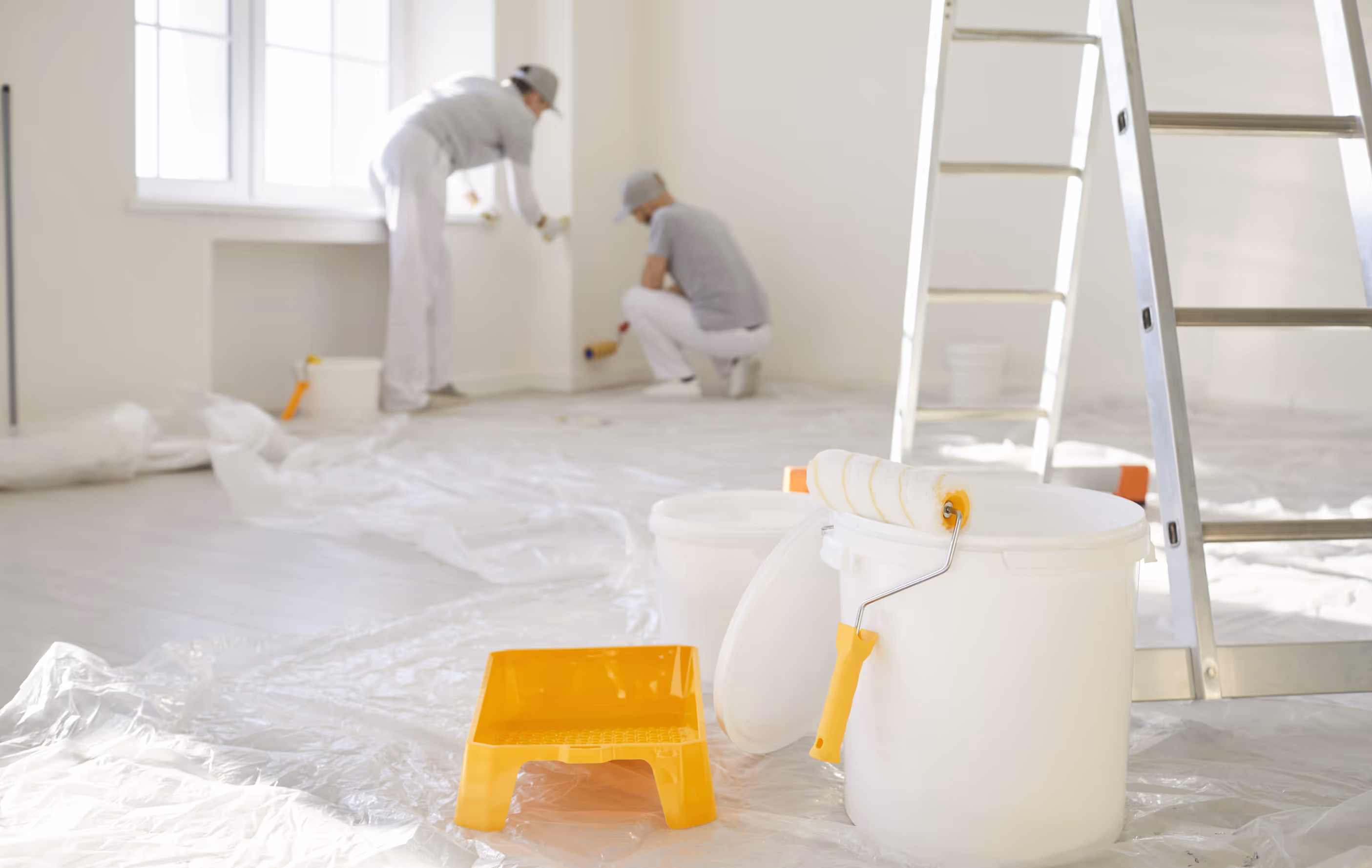 Two painters in white and gray clothes painting a white room with a paint roller near a window, with painting supplies and a ladder in the foreground.