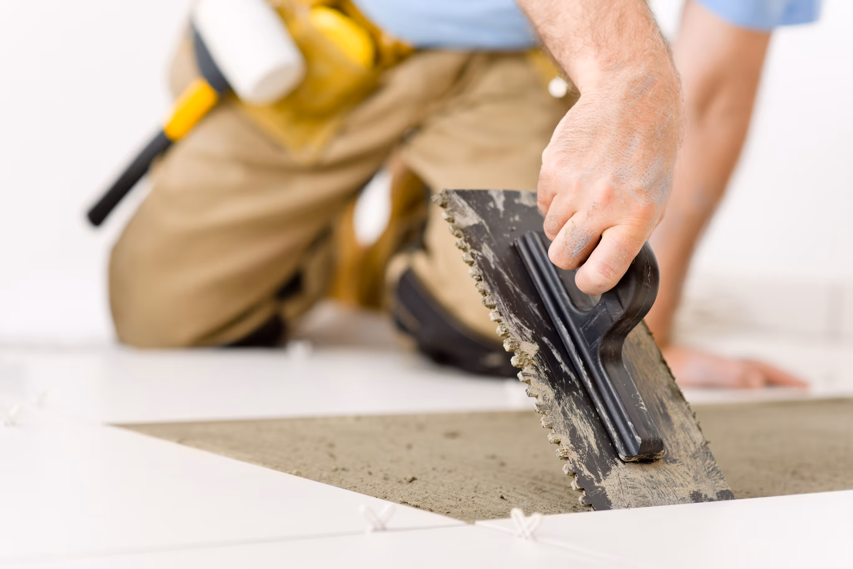 Worker spreading adhesive with a notched trowel while installing white floor tiles.
