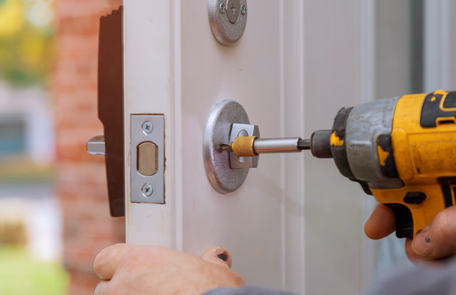 Close-up of a hand using a yellow cordless drill to fix or install a lock on a white door.
