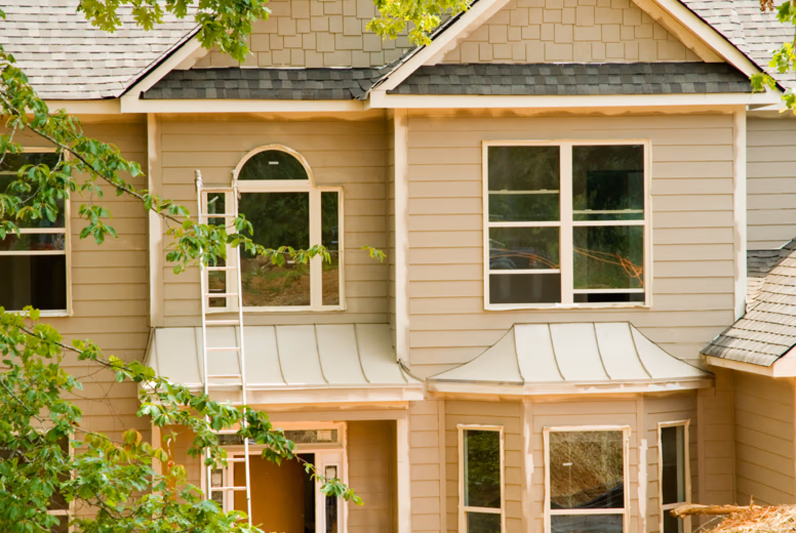 Beige house exterior with multiple windows, a ladder leaning against the wall, and green tree branches partially covering the view.