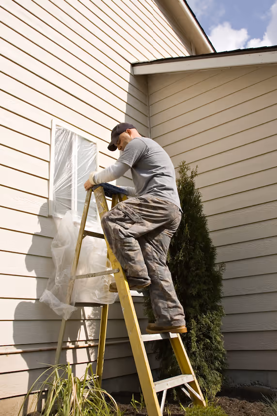 Man in a cap and paint-splattered pants standing on a yellow ladder, preparing to paint the exterior wall of a house with plastic-covered window.