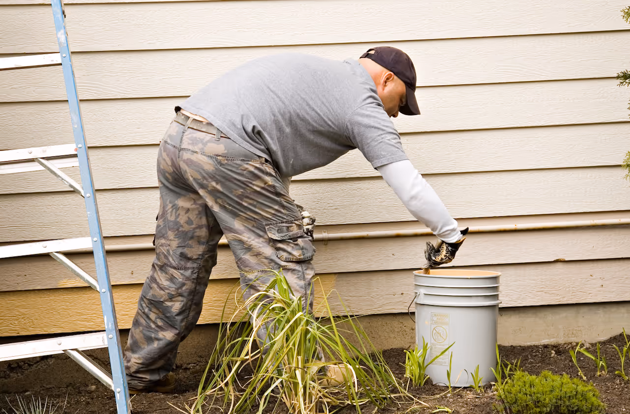 Man in camouflage pants and gray shirt stirring paint in a large gray bucket next to a house siding and a blue ladder.
