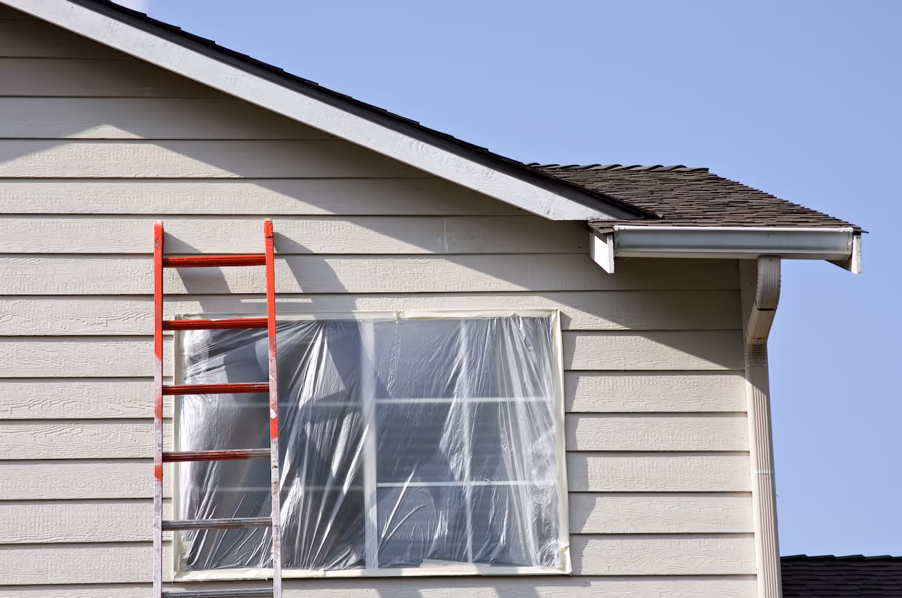 Red ladder leaning against a beige house with a plastic-covered window under clear blue sky.