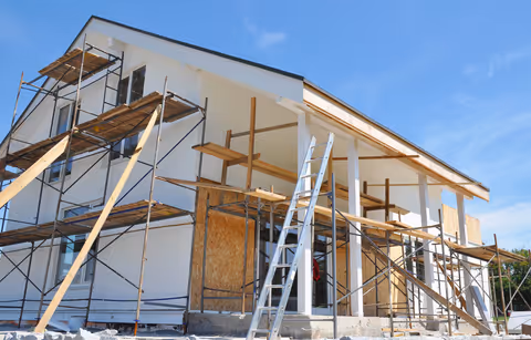 House under construction with scaffolding and ladders against a clear blue sky.