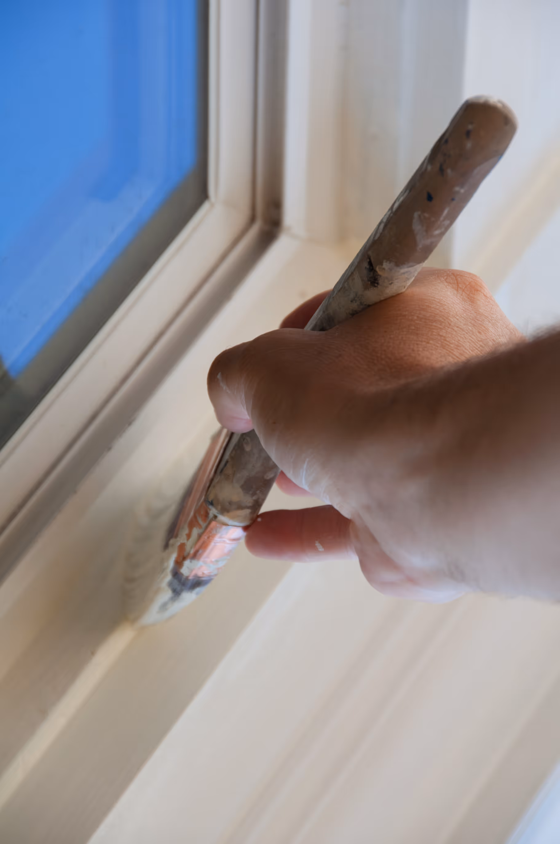 Hand painting a white window frame with a paintbrush near blue glass.
