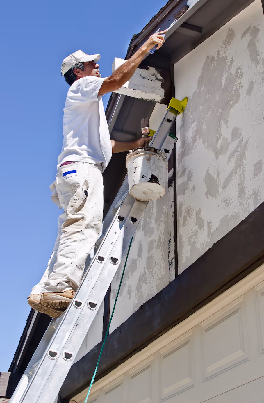 Man in white clothes painting the exterior upper wall of a house while standing on a ladder under clear blue sky.