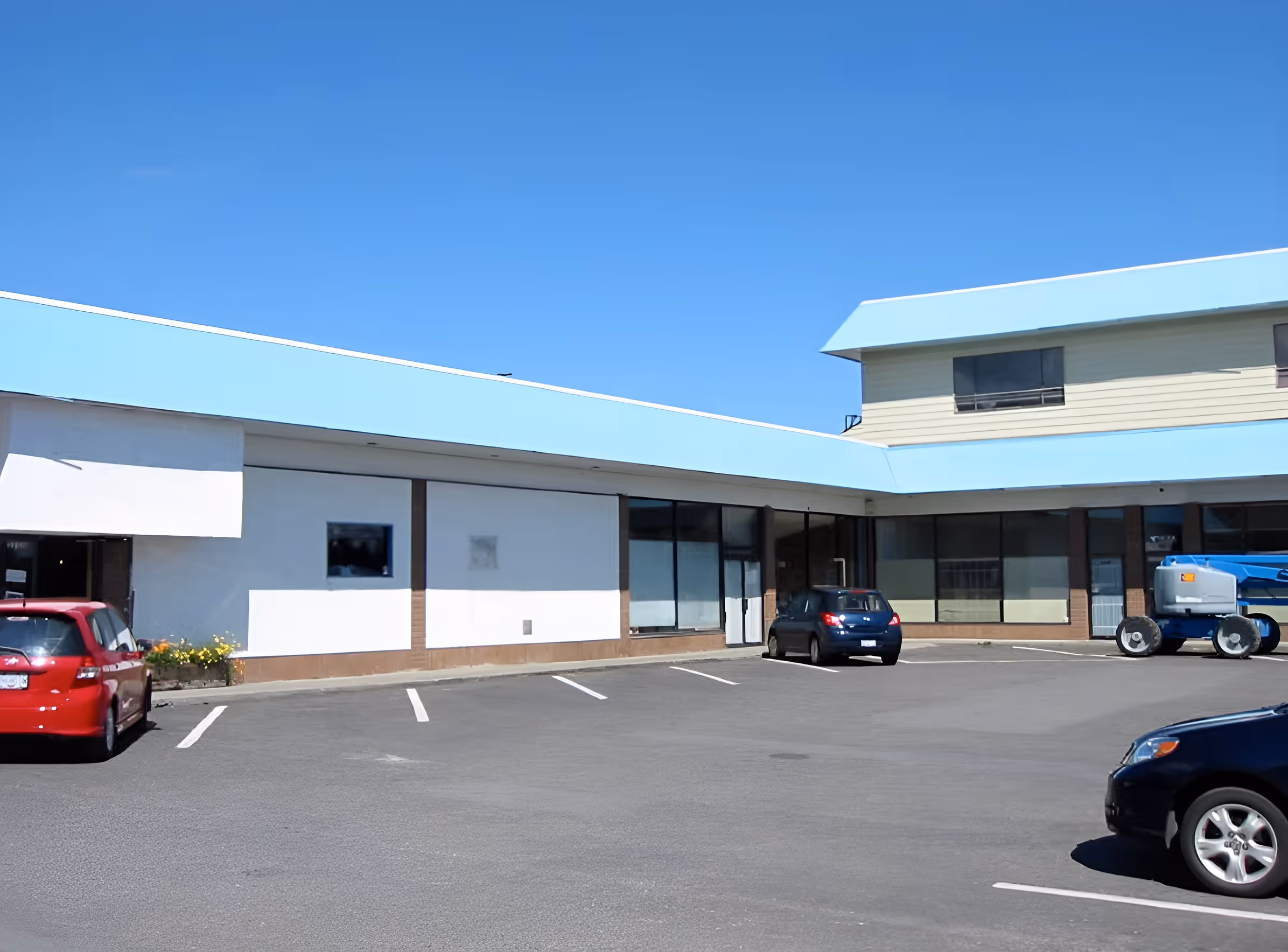 Parking lot with a few cars in front of a building with light blue roof and large windows under a clear blue sky.
