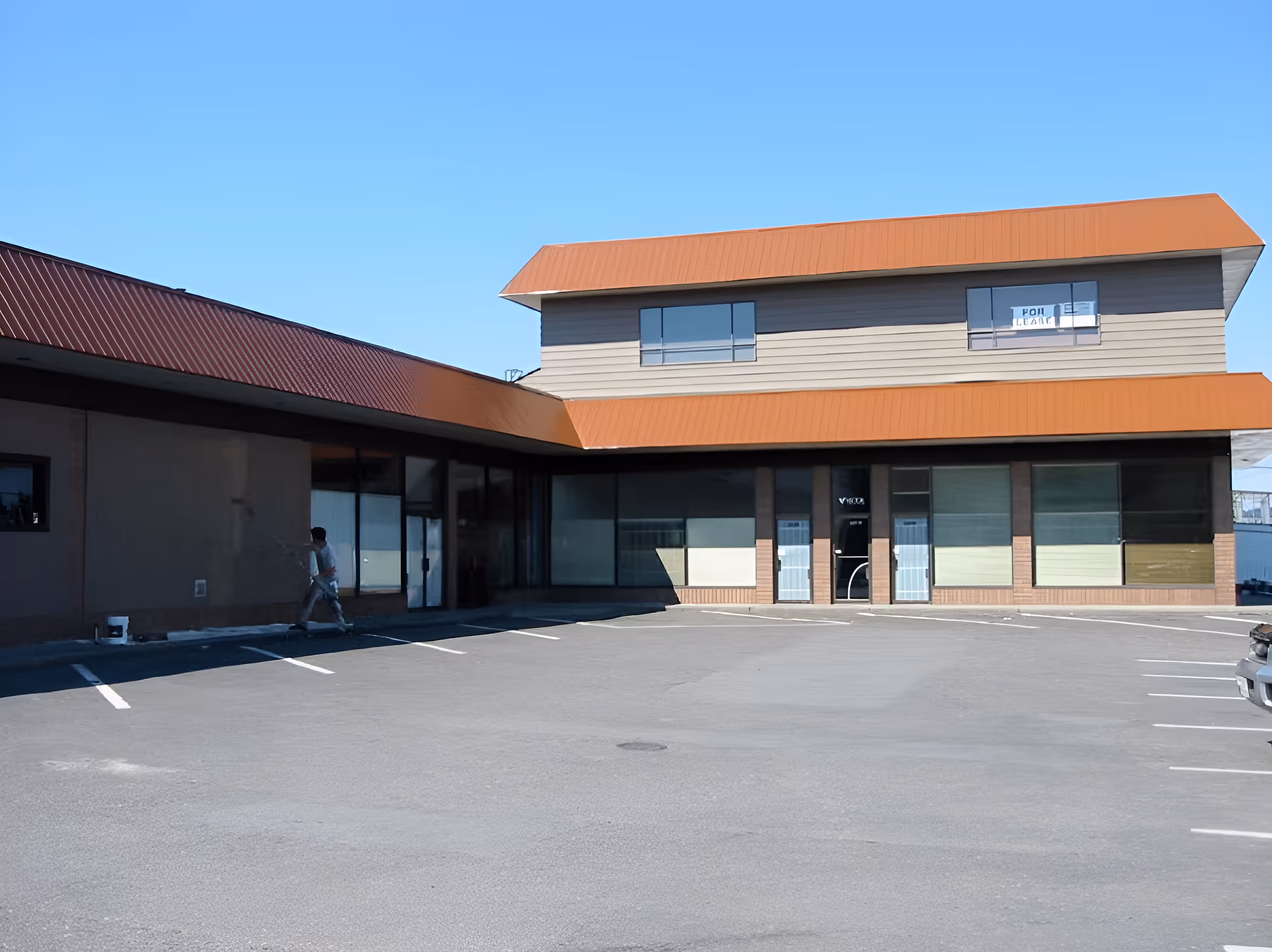 Empty parking lot in front of a two-story commercial building with orange roofs under a clear blue sky.
