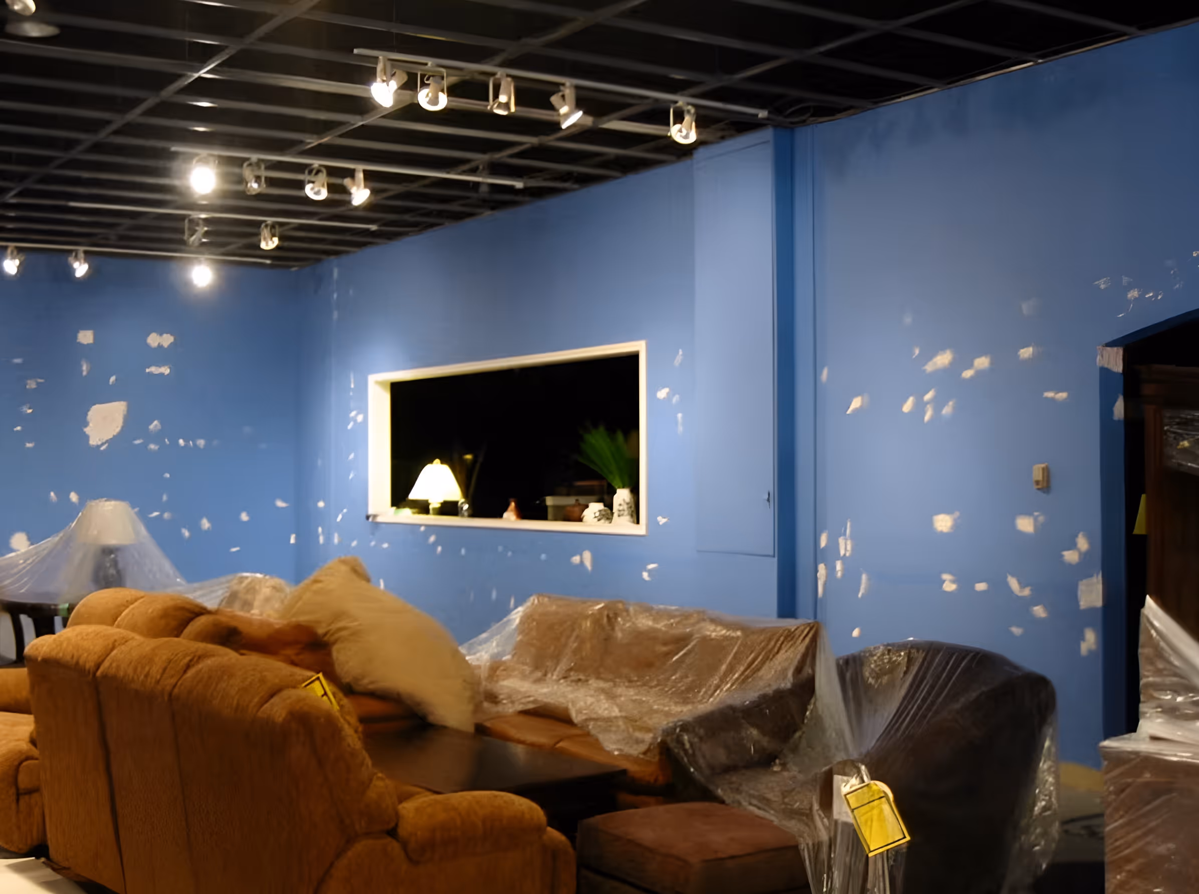 Living room with blue walls showing patch repairs, brown sofa set and armchair covered in plastic, and ceiling lights.