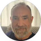 Older man with gray hair and beard smiling indoors by a window with sheer curtains.