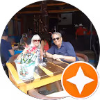 Two people wearing sunglasses sitting at an outdoor table with a flower vase and menus, under a covered patio.