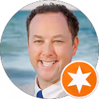 Smiling man with short brown hair wearing a suit and blue tie against a blurred outdoor background.