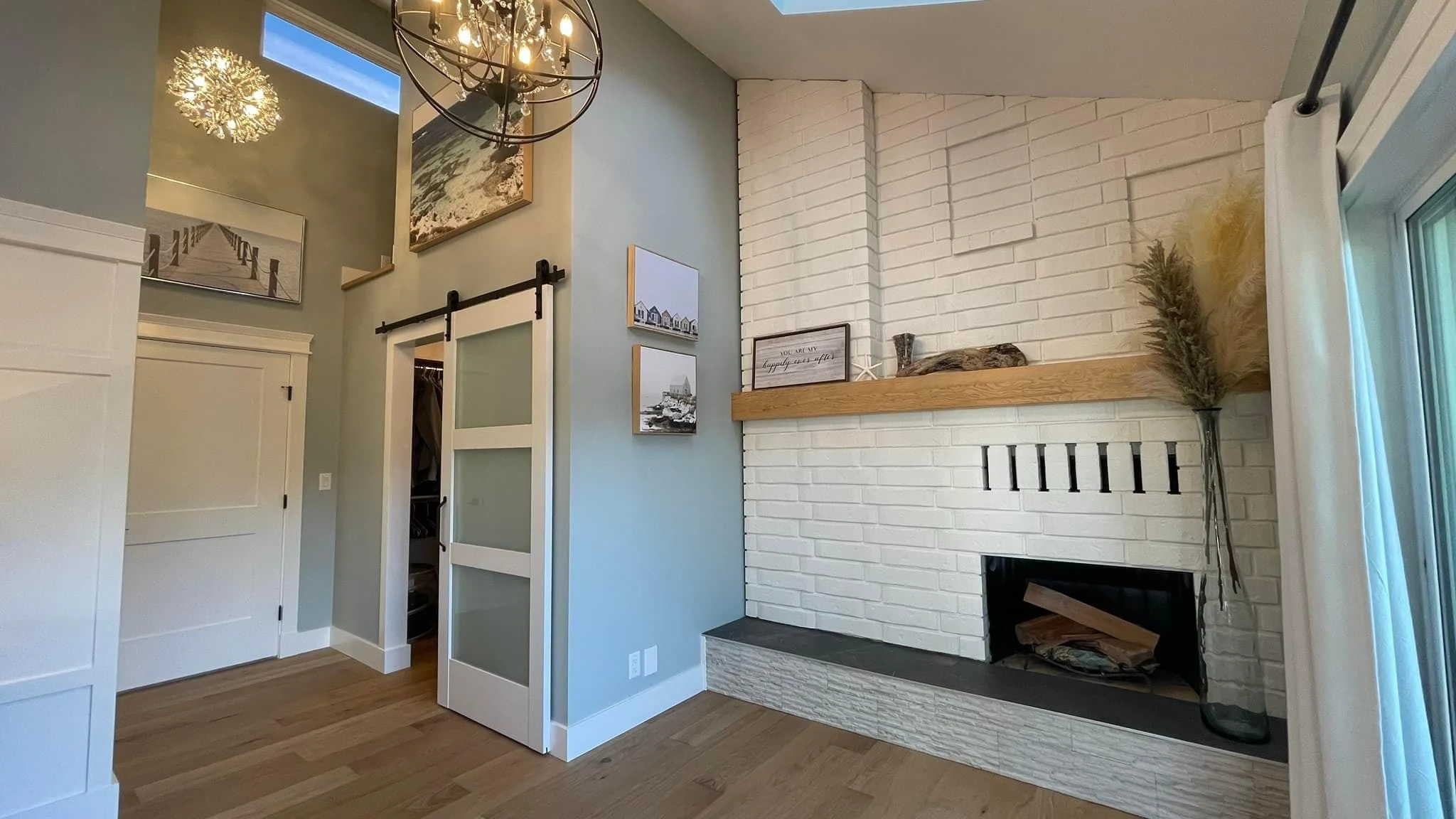 Modern corner room with white brick fireplace, wooden mantel with decorative items, blue-gray walls, sliding barn door, and light wooden floor.