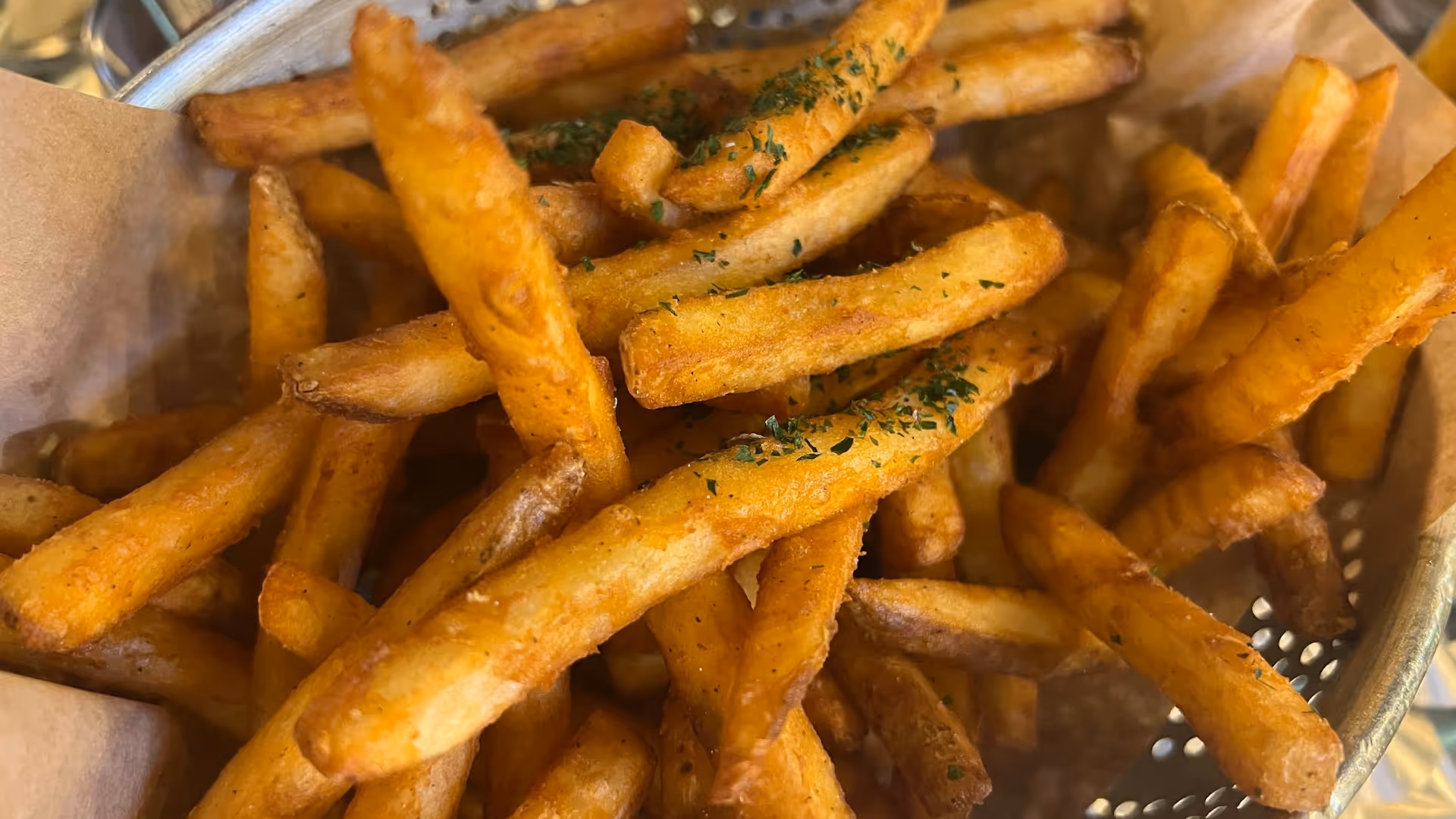 Close-up of golden French fries sprinkled with green herbs in a metal basket.
