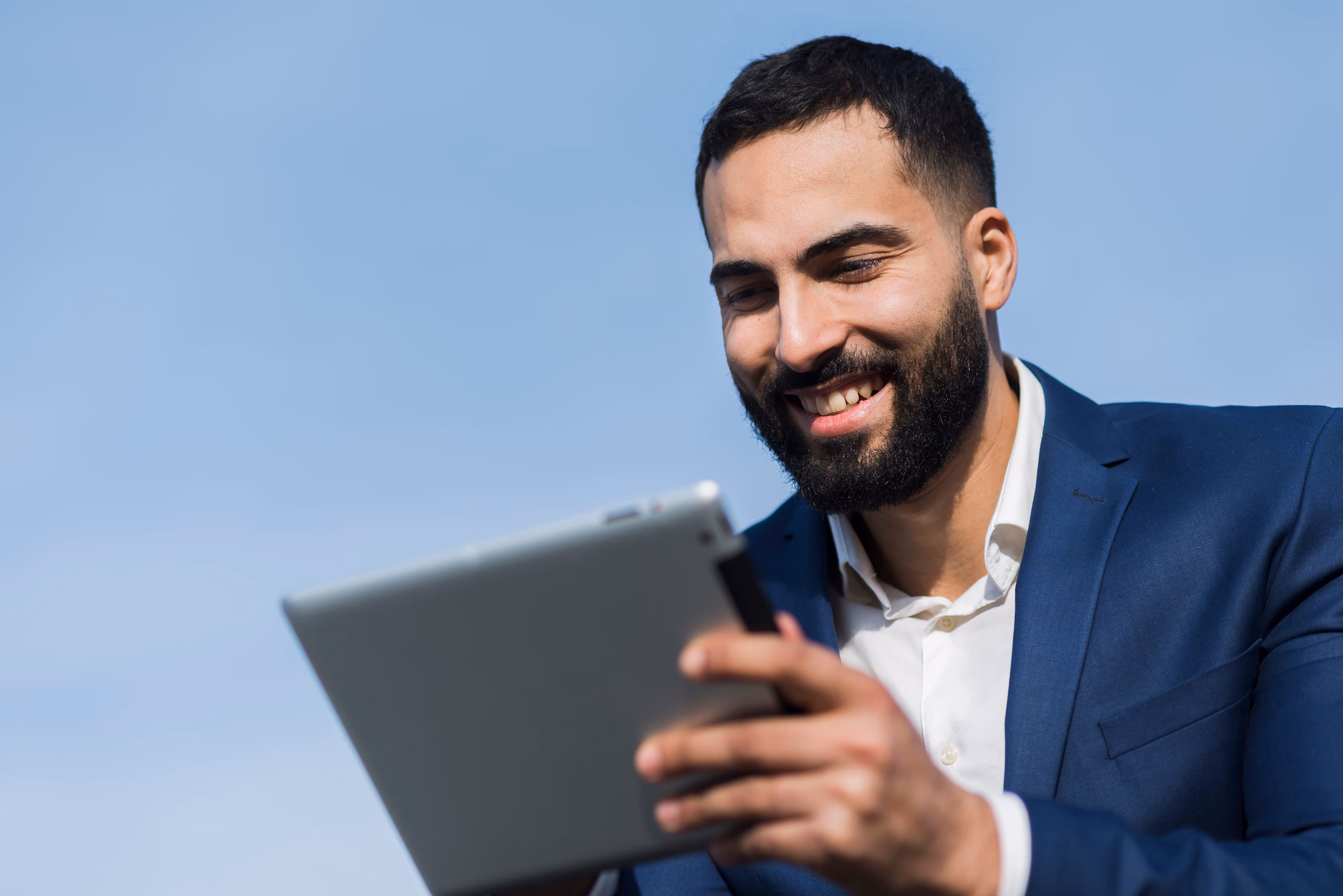 Smiling man with beard in a blue suit using a tablet outdoors against a clear sky.